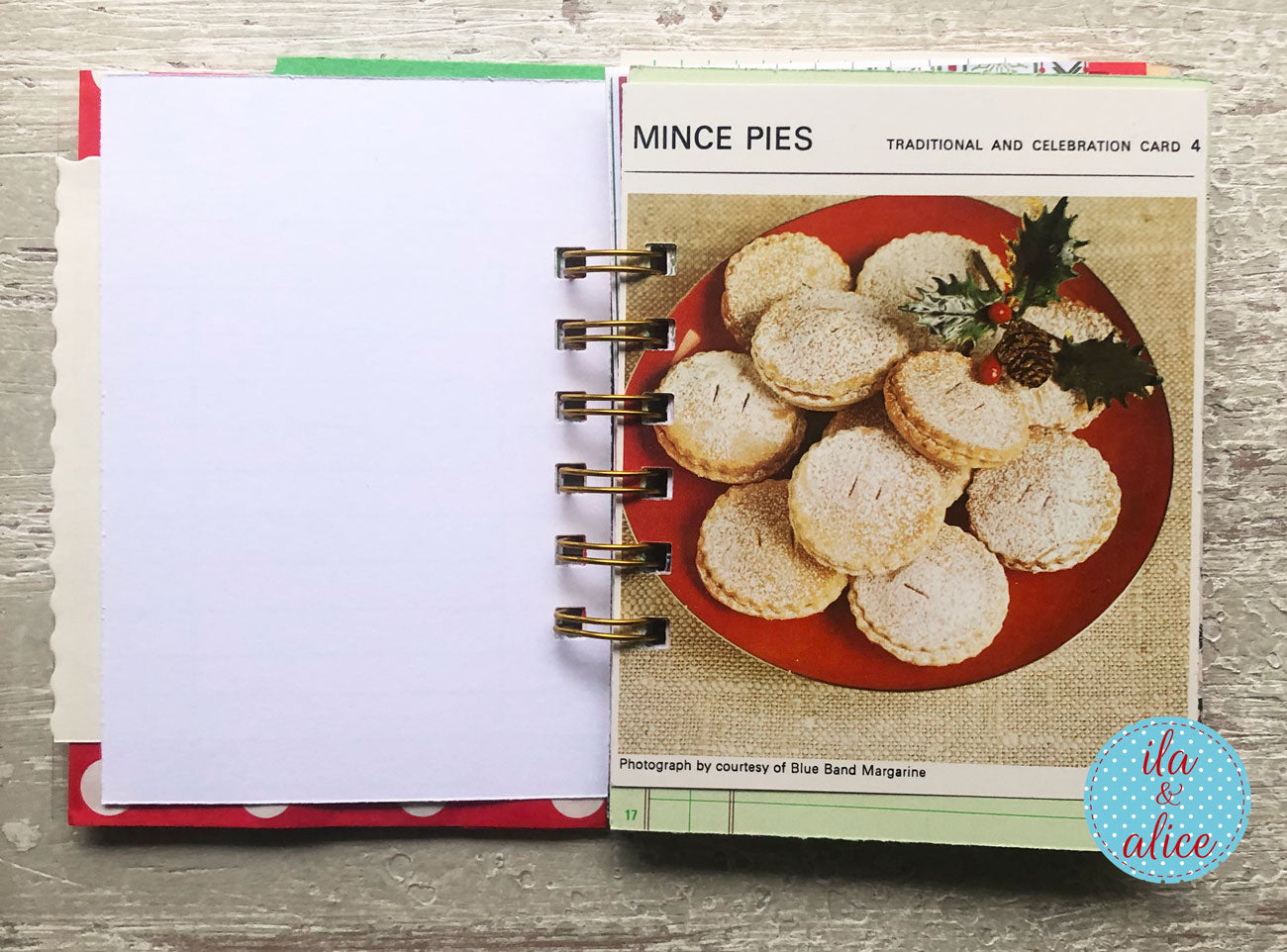 An open spiral-bound recipe or card book on a gray wood surface, showing a page titled 'MINCE PIES' with a photo of powdered sugar-coated mince pies on a red plate. The left page is blank white, and a blue circular 'ila & alice' watermark appears in the lower right corner.