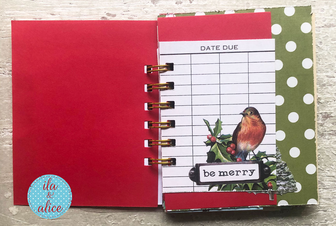 A handmade spiral-bound scrapbook journal or planner page set on a light wood background. The pages feature festive holiday designs, including a red cover, a green and white polka dot page, a bird and holly graphic with the text 'be merry', and a date due grid.