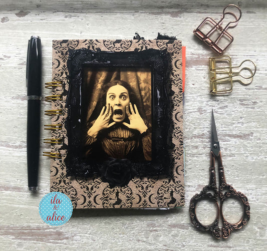 A handmade spiral-bound journal with a gothic damask pattern cover, black lace trim, and a central sepia-toned portrait of a shocked woman. The journal is styled on a whitewashed wooden surface alongside a black pen, metal paperclips, and ornate scissors.