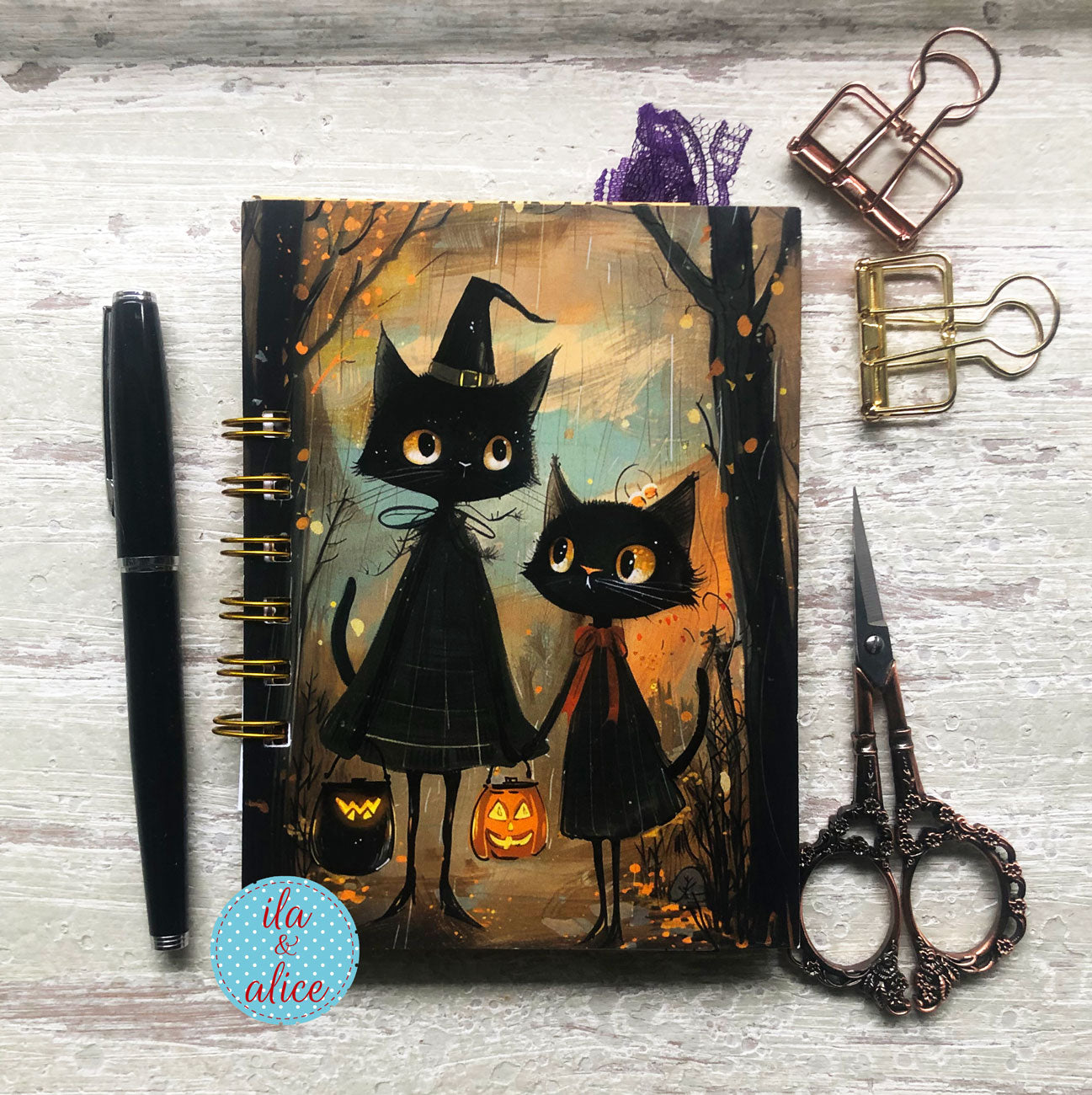 A spiral-bound planner or notebook with a Halloween illustration of two black cats in witch hats, styled on a whitewashed wood surface with a black pen, metal paperclips, ornate scissors, and a blue circular sticker.