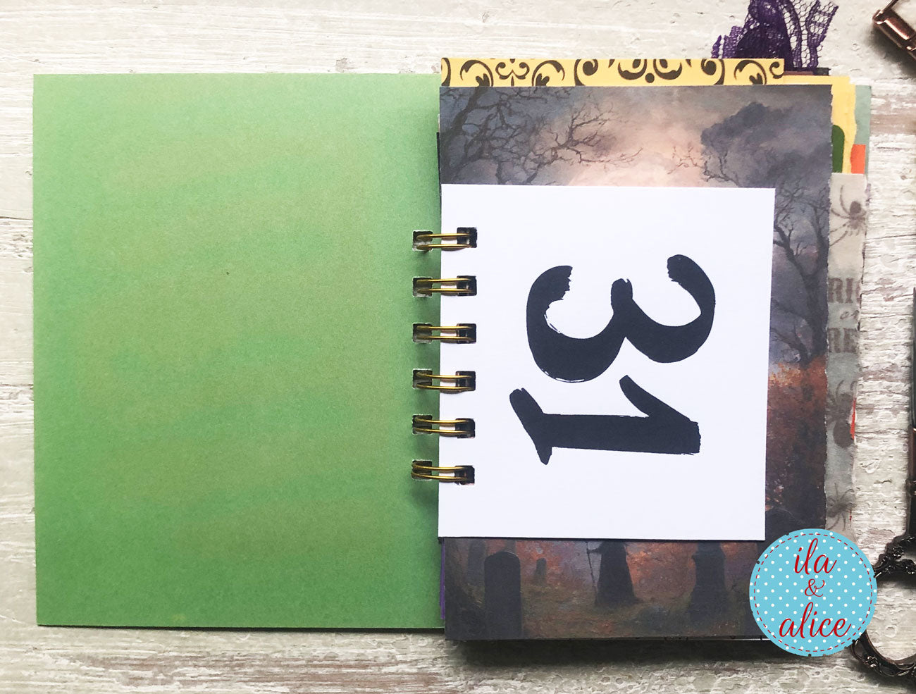 A spiral-bound notebook or journal laid open on a whitewashed wood surface. The left page is a solid green cover, and the open middle section shows a white page with the number 31 in black script, layered over a spooky graveyard photo page.