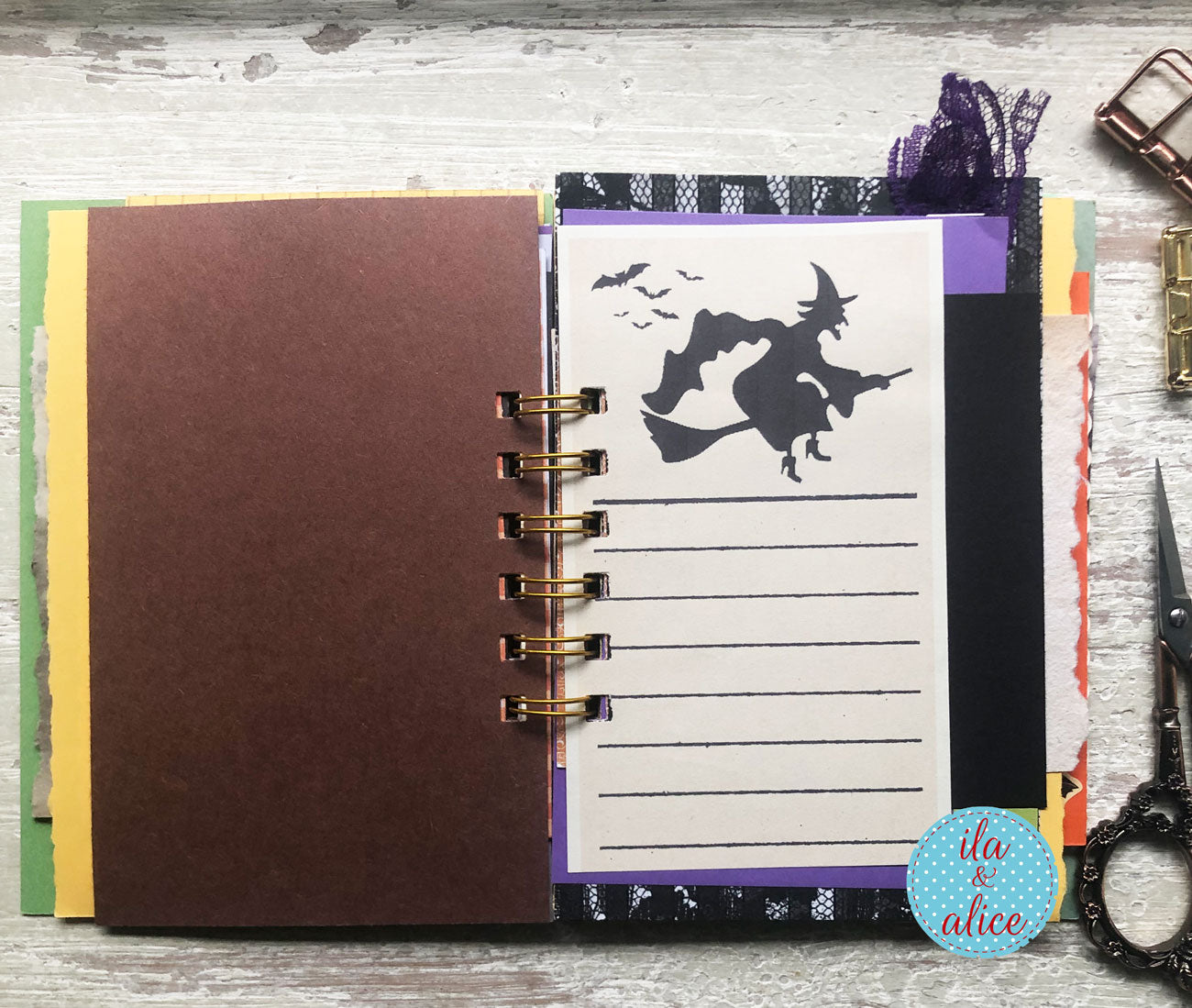 A handmade spiral-bound journal or notebook shown open on a whitewashed wooden surface. The left page is a large dark brown card, and the right page features a white lined writing area with a black witch silhouette and bats, bordered by patterned strips in purple, black, and white.