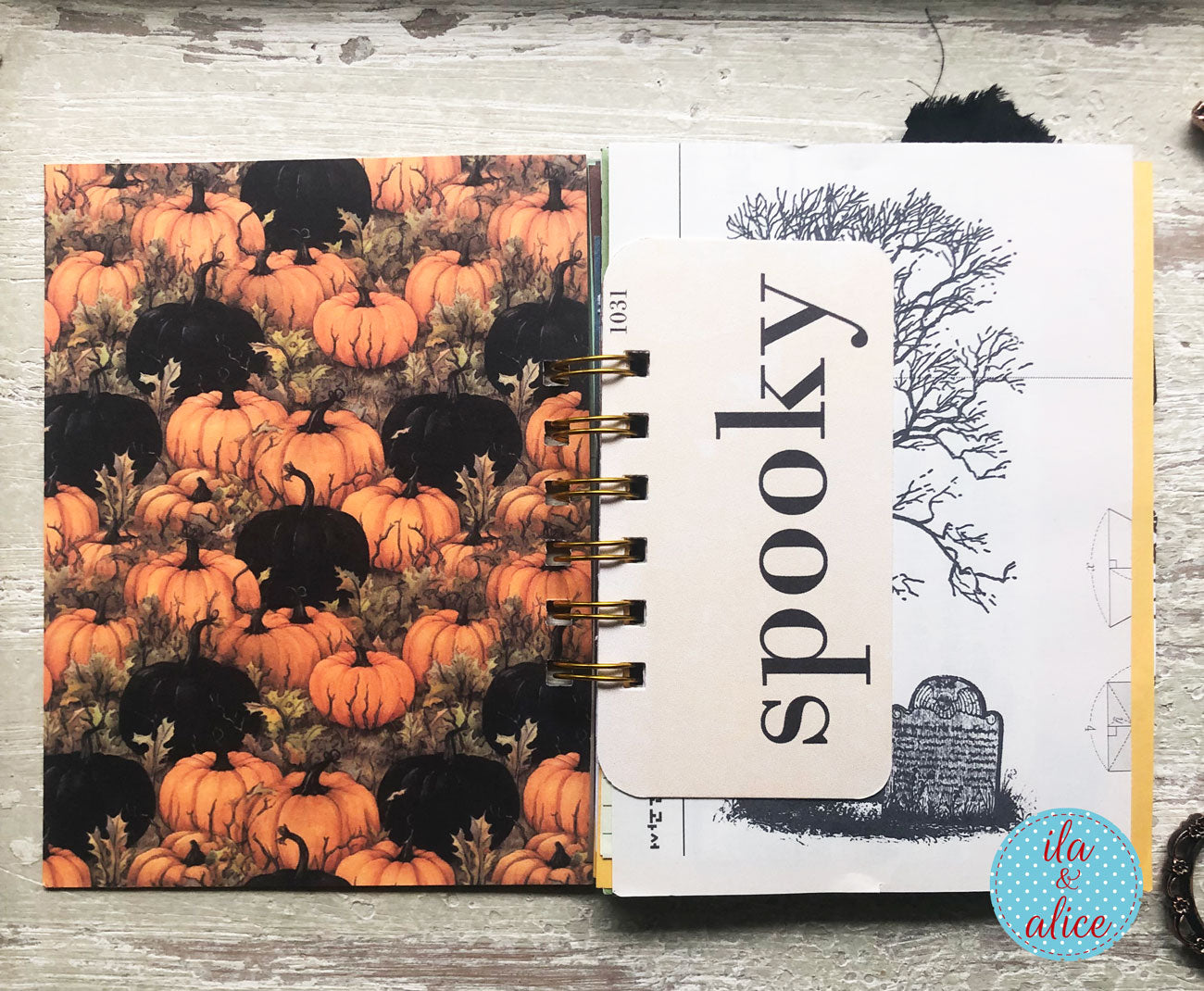 An open spiral-bound notebook or journal laid flat on a distressed white wood surface. The left page shows a dense pumpkin and black cat pattern, and the right page has a tombstone illustration with the word 'spooky' on a white card overlay.
