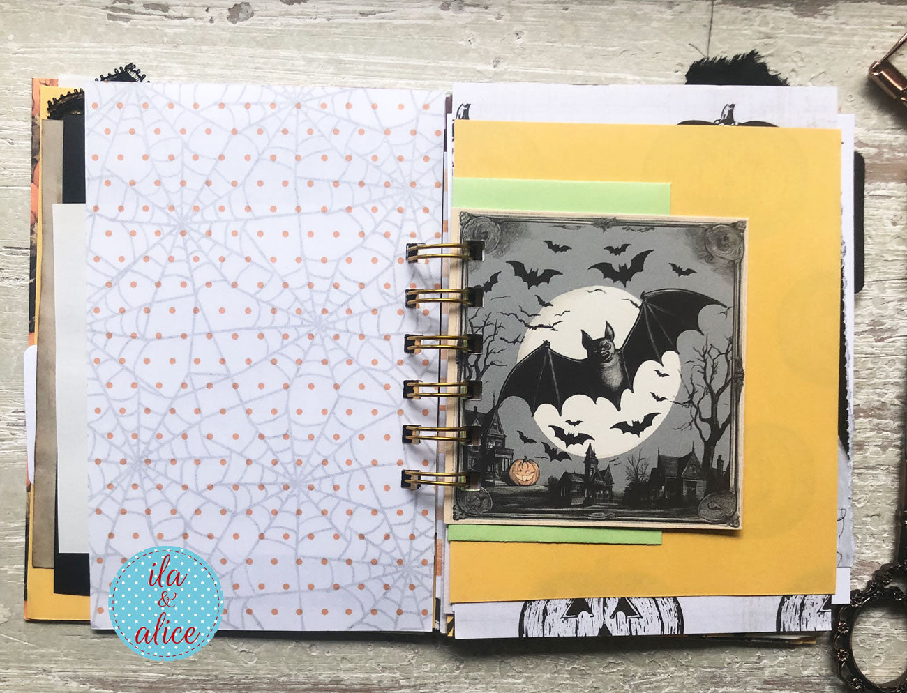 An open spiral-bound scrapbook journal or art notebook on a rustic wood surface, featuring layered Halloween-themed paper elements including a gray bat collage on yellow cardstock, patterned white paper with orange dots, and a circular blue 'ita alice' sticker.