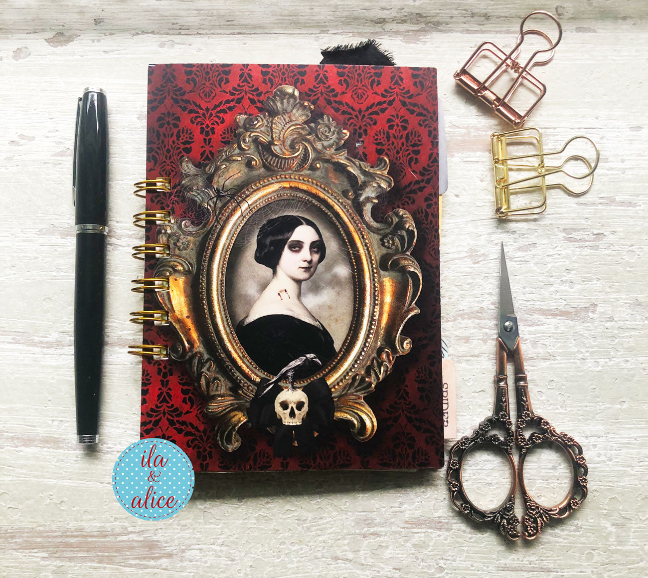 A gothic-style spiral notebook with a red damask cover, featuring an ornate gold frame illustration of a woman's portrait, a skull charm, and a black feather accent. The notebook is styled in a flat-lay on a light wood background alongside a black pen, gold and rose gold binder clips, and antique-style scissors.