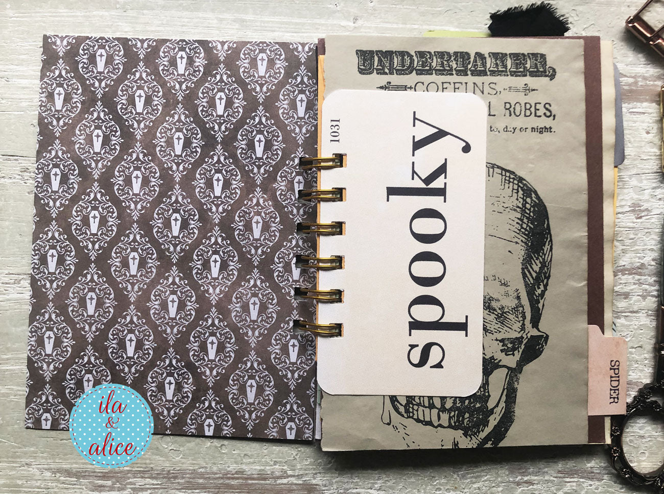 An open spiral-bound notebook or journal laid flat on a rustic wood surface. The left page has a dark brown and gray damask pattern with small tuxedo silhouettes.