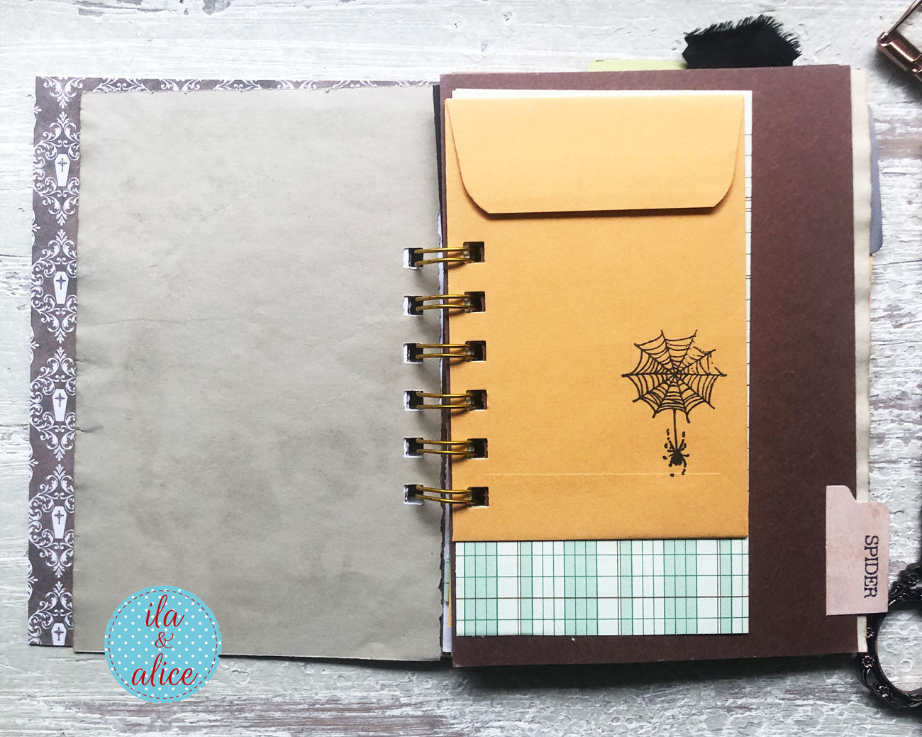 A handmade spiral-bound planner or notebook shown open on a whitewashed wood surface. The left page is a textured gray card with a patterned gray-and-white border.