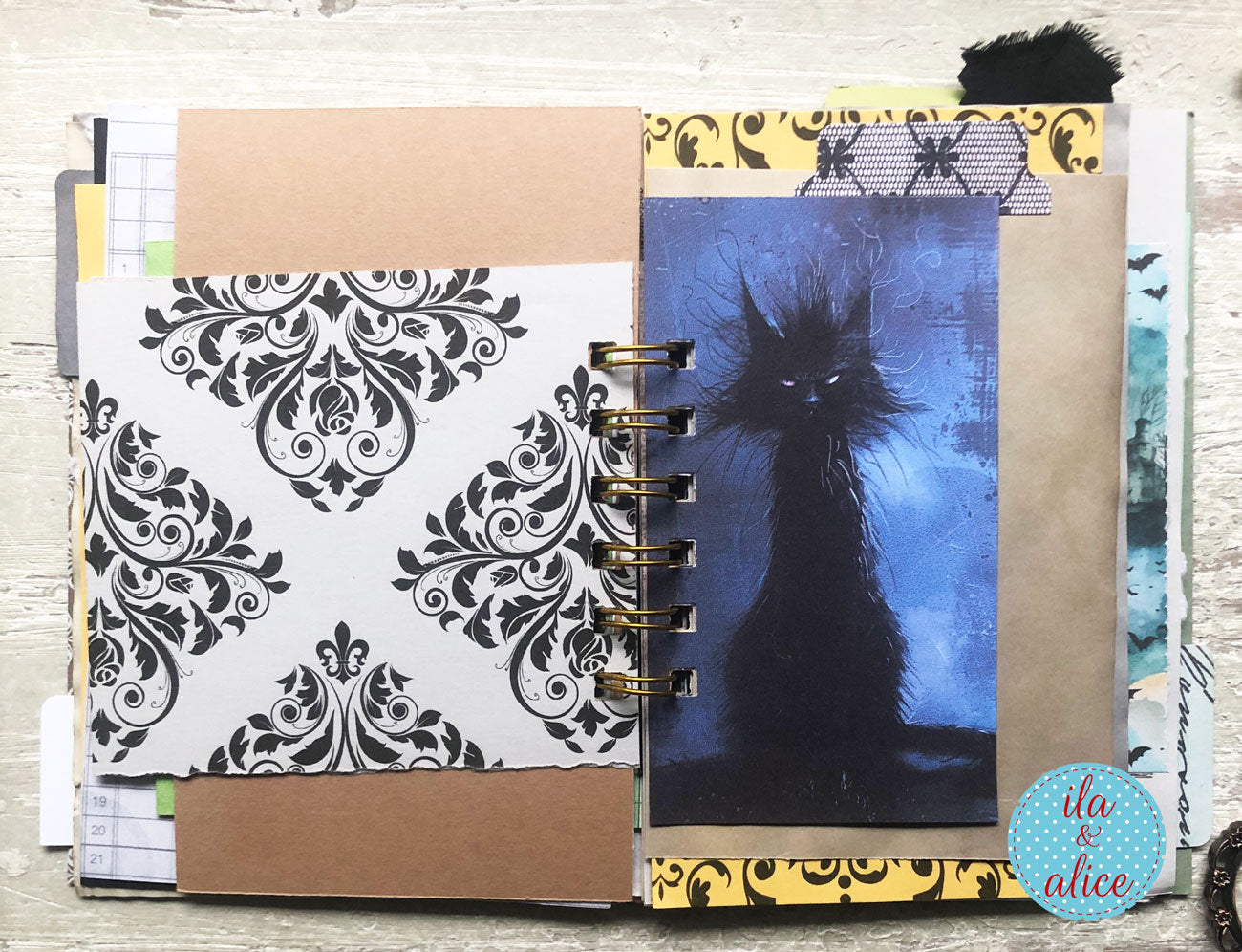 A handmade spiral-bound scrapbook journal or art book shown open on a light wood surface. The pages feature black-and-white damask patterned paper, kraft brown paper tabs, yellow patterned borders, and a central blue-toned illustration of a black cat.