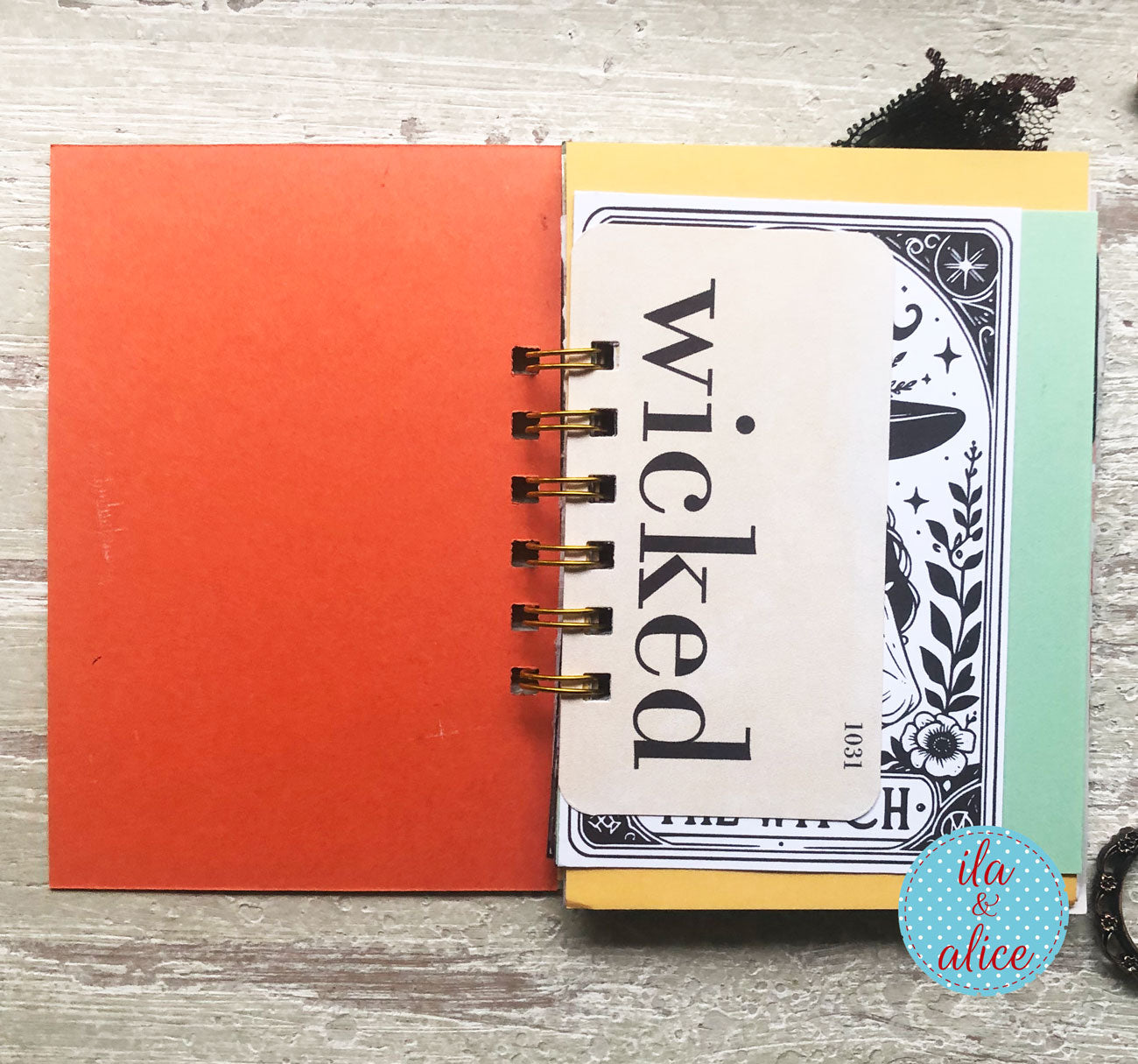 A handmade spiral-bound notebook or journal featuring a vintage-style card cover with the word 'wicked' in black script, layered over a bright coral-orange sheet and a mint green sheet. The notebook is photographed from above on a light gray wood-textured surface with a blue circular 'ita & alice' logo sticker in the bottom right corner.