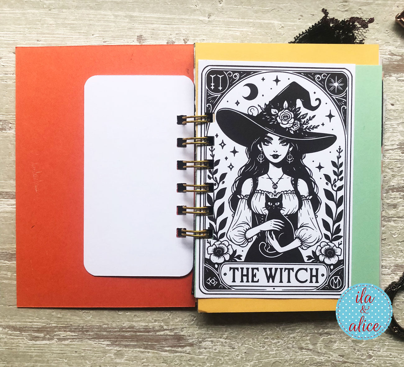 A spiral-bound notebook or planner page set displayed on a light wood surface. The right page features a black-and-white tarot-style illustration of 'The Witch' wearing a large hat with flowers, surrounded by decorative line art.