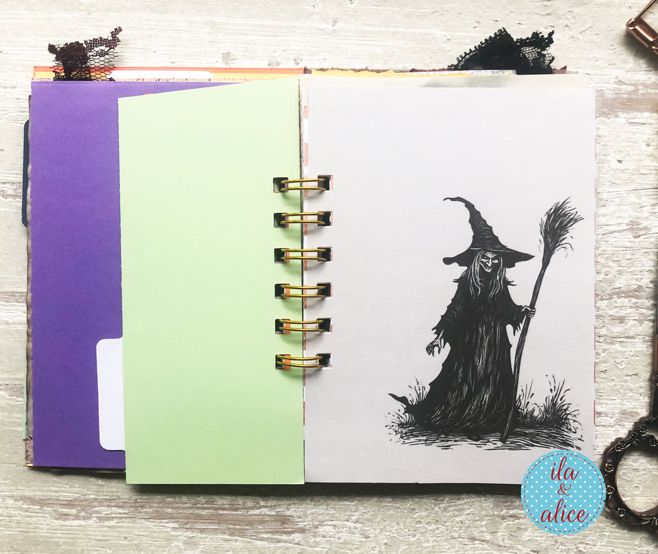 An open spiral-bound notebook or journal lying on a light wood surface, showing a black ink illustration of a witch holding a broom on the right page. The left page is pale green, and a purple sheet is visible underneath.