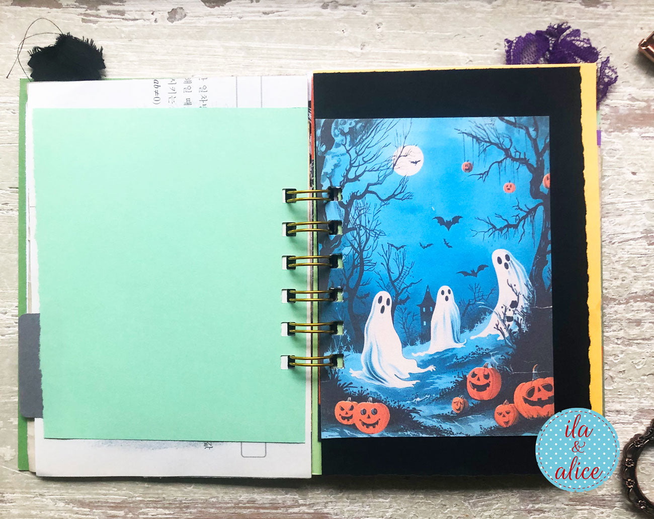 An open handmade spiral-bound journal on a rustic wooden table, featuring a colorful Halloween-themed illustration of three ghosts, pumpkins, and bats on the right page, alongside a blank mint green paper sheet on the left.