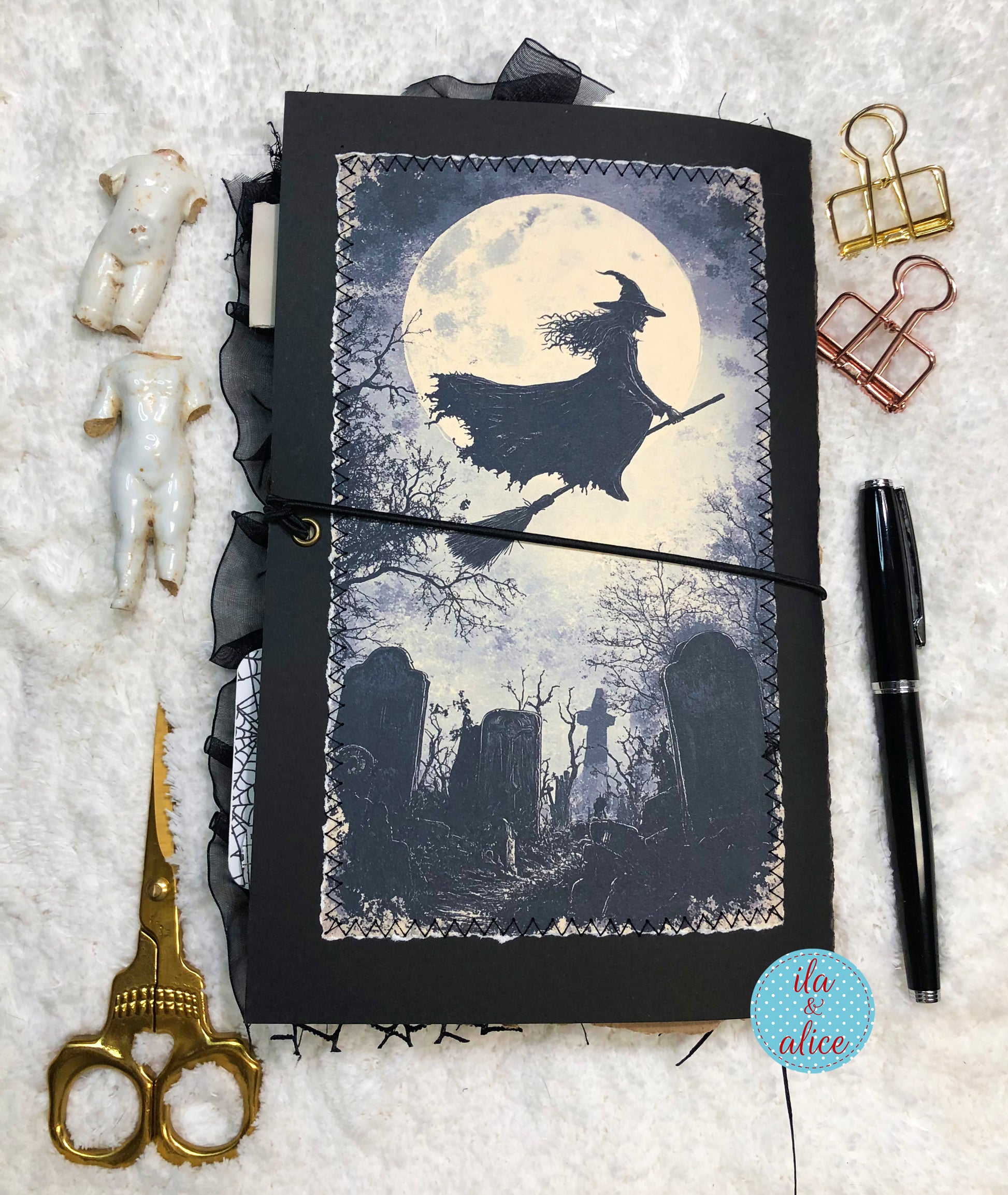A handmade witch-themed journal with a black cover and a printed illustration of a witch flying on a broomstick in front of a full moon over a graveyard, styled on a white textured background with gold scissors, white figurines, binder clips, a black pen, and a blue 'do & alice' sticker.