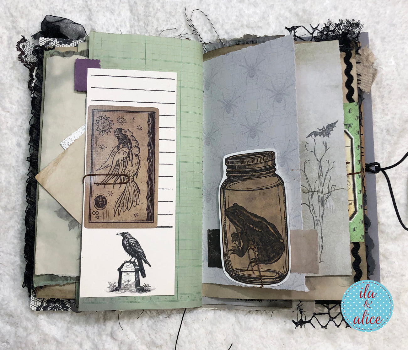 An open handmade mixed-media journal spread on a white textured fabric background. The page features layered paper elements including sage green lined paper, gray spider-patterned paper, black lace trim, a vintage-style bird illustration card, a frog-in-a-mason-jar sticker, and other collage elements.