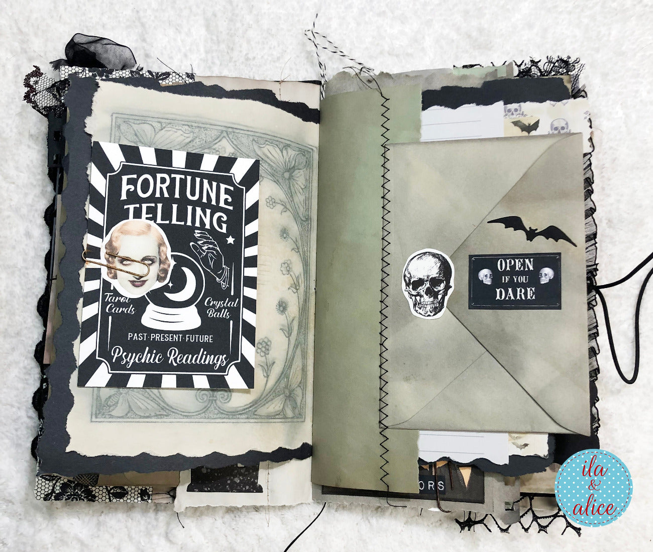An open handmade gothic-themed mixed-media journal spread, featuring distressed black and white pages with a 'Fortune Telling' card insert, a skull sticker, a black envelope pocket labeled 'Open to the Dark', and a small bat sticker, all laid flat on a white textured fabric background.