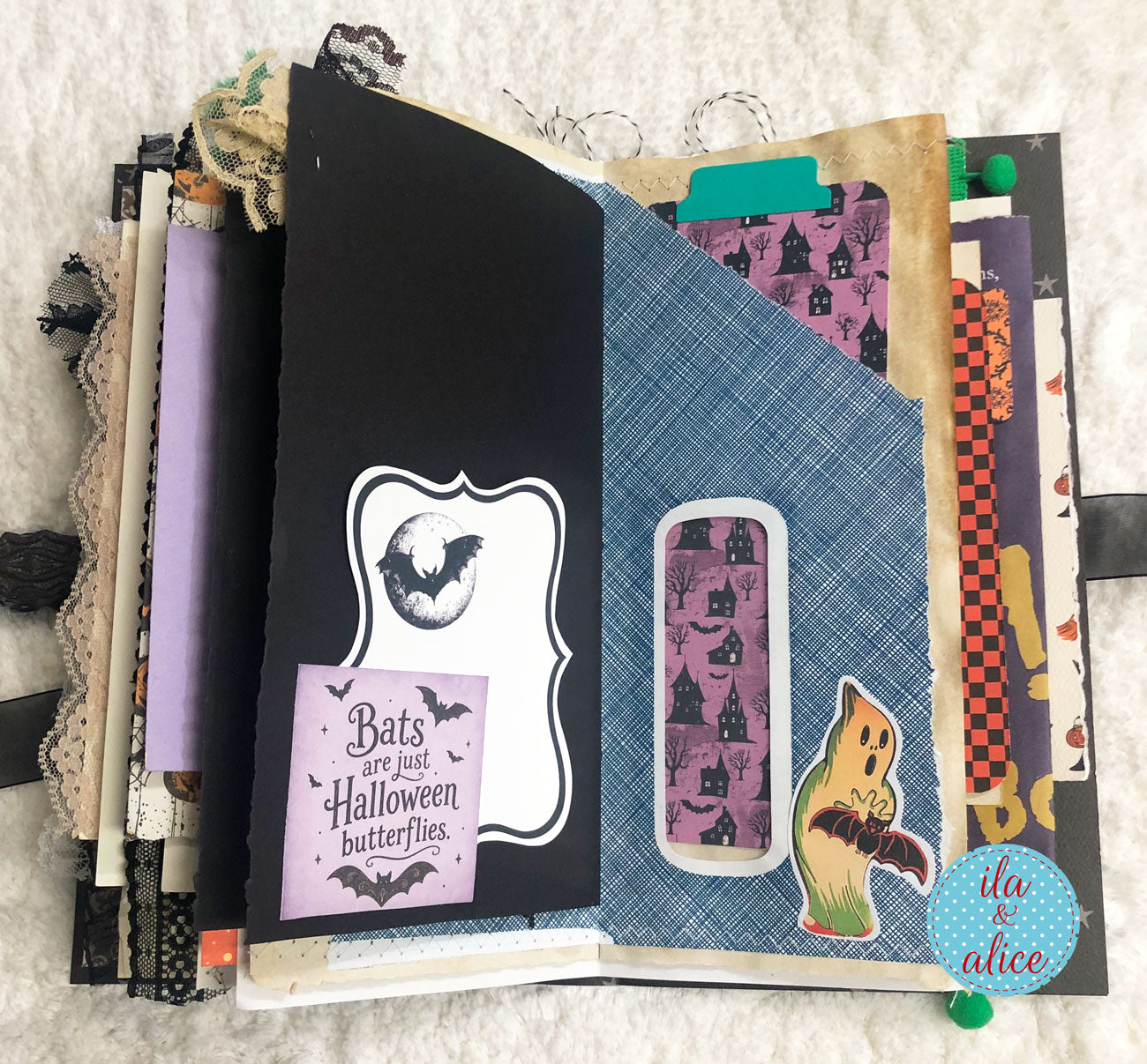 A handmade Halloween-themed scrapbook album or journal spread open on a white textured fabric background. The pages feature black, blue, purple, and brown elements, including patterned paper with bats and cats, a die-cut sticker of a ghost, and decorative tabs.