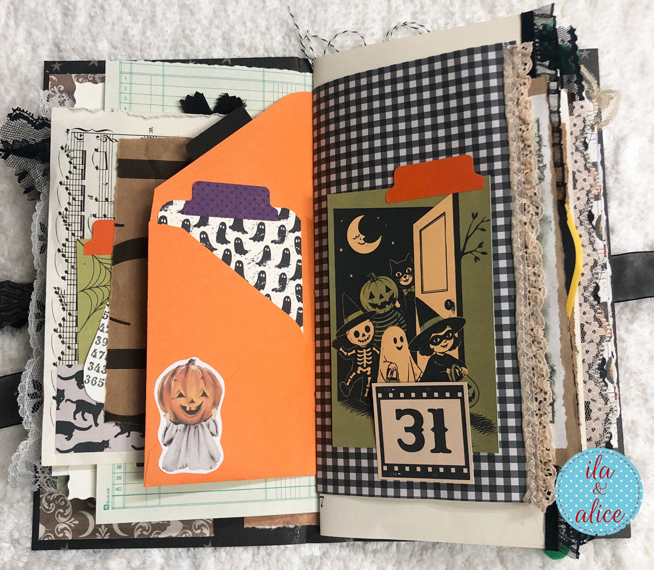 An open handmade scrapbook journal or art book displayed on a white textured fabric background. The pages feature Halloween-themed decorations including an orange pocket fold with a jack-o'-lantern sticker, black-and-white gingham paper, lace embellishments, musical notes, and a date tab showing '31'.