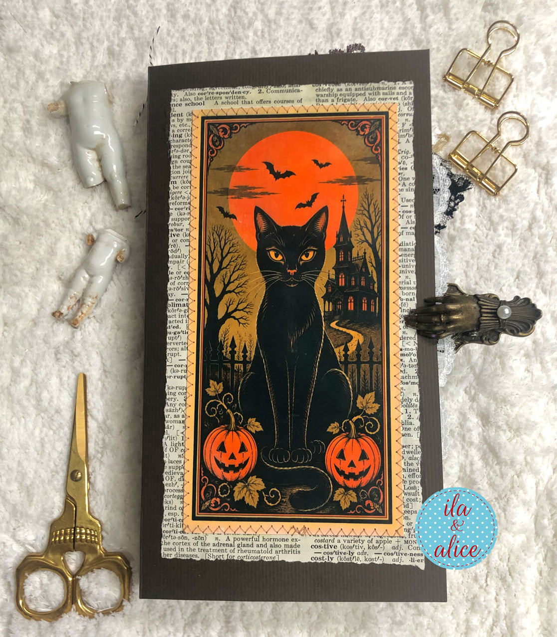 A Halloween-themed art print or card featuring a black cat in front of an orange full moon, displayed on a textured white fabric background with decorative scissors, binder clips, and small figurines.