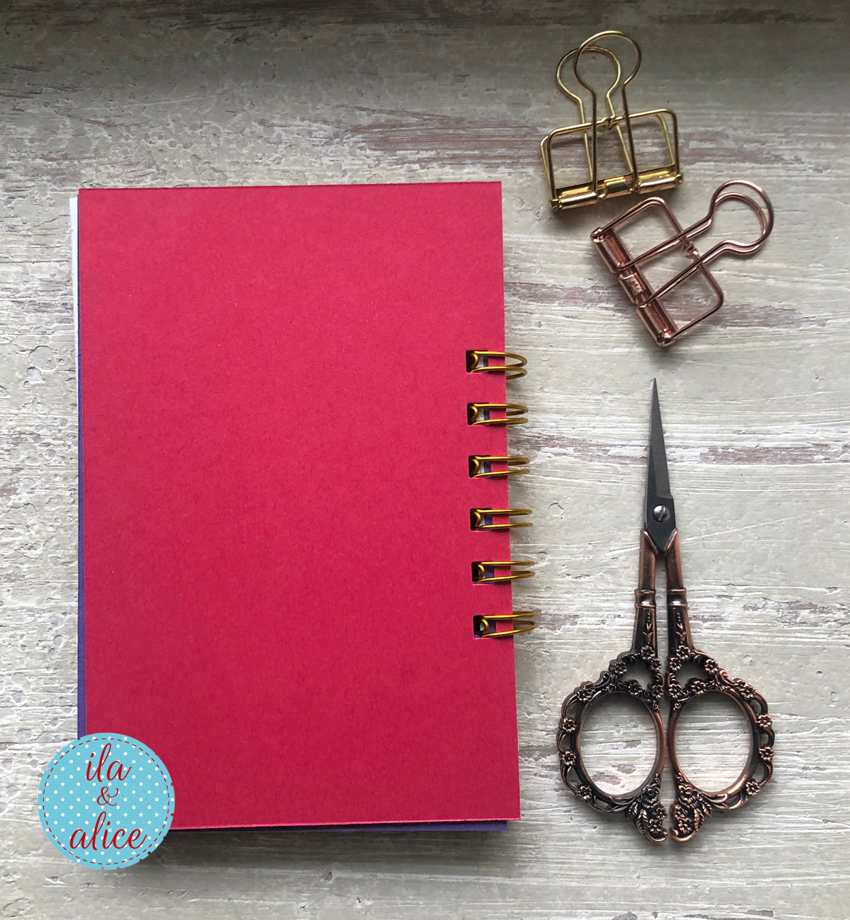 Red notebook with gold binding, vintage scissors, and paper clips on a wooden surface.