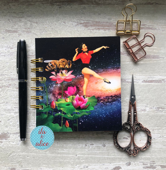 Notebook with a colorful design featuring a woman, bee, and lotus flowers on a wooden surface with stationery items.