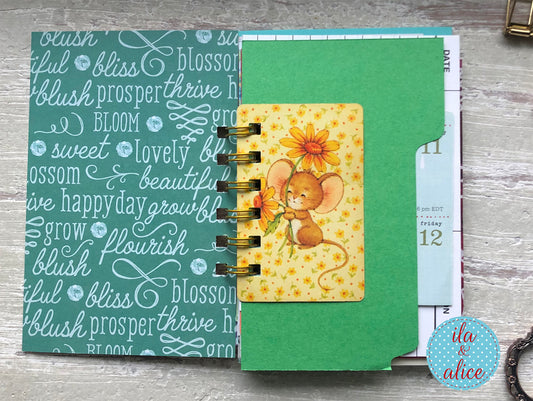 Small spiral-bound notebook with a decorative cover featuring a mouse and flowers on a wooden surface.
