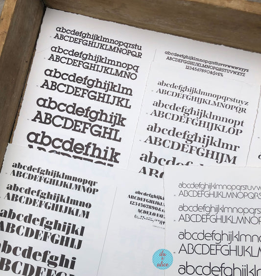 A close-up of printed alphabet lettering sheets showing uppercase and lowercase serif font samples arranged inside a wooden tray or box.